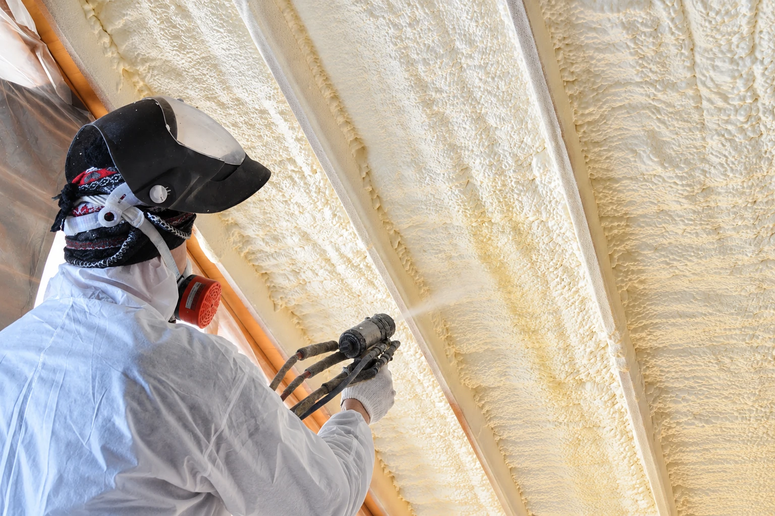 Worker in protective gear sprays foam insulation between overhead wooden rafters.