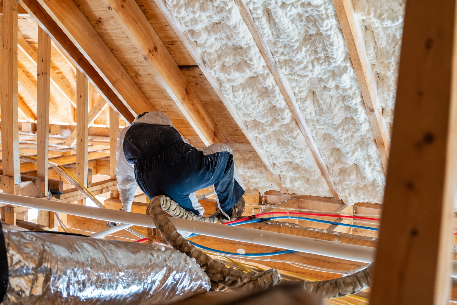 A worker applies white spray foam between sloped attic rafters in a timber-framed space.