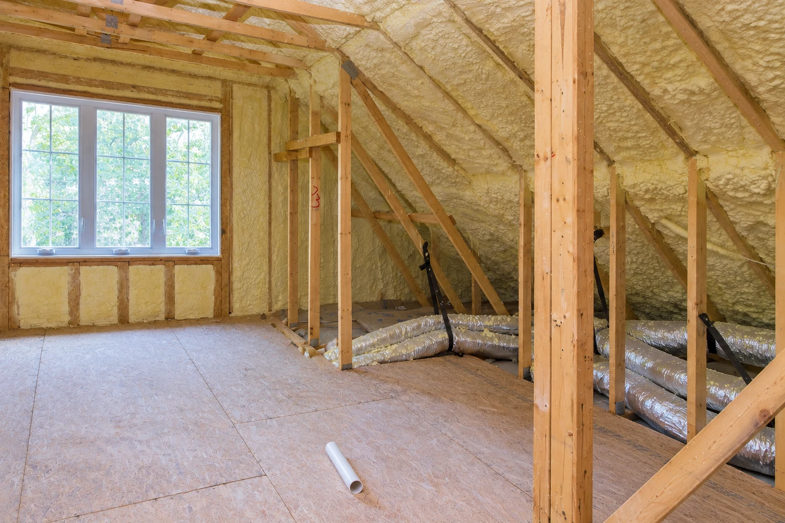 Attic room with exposed wooden framing, spray foam coating the sloped ceiling and walls, and a large window.