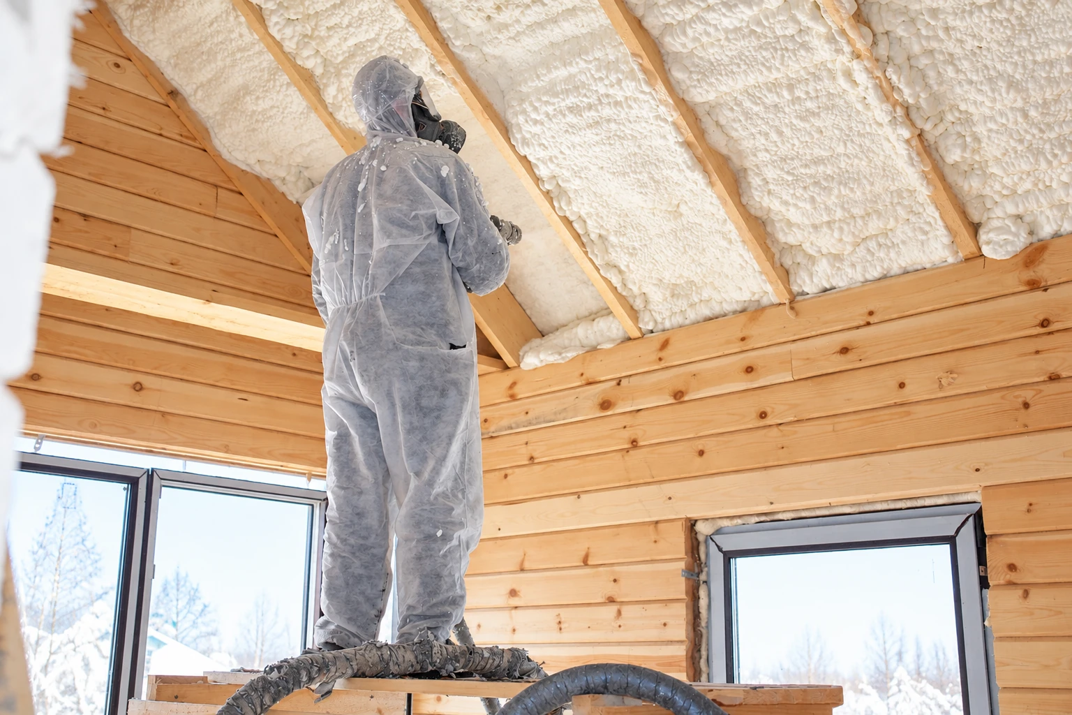 A worker in protective coveralls sprays white foam insulation between the exposed rafters of a wooden ceiling.