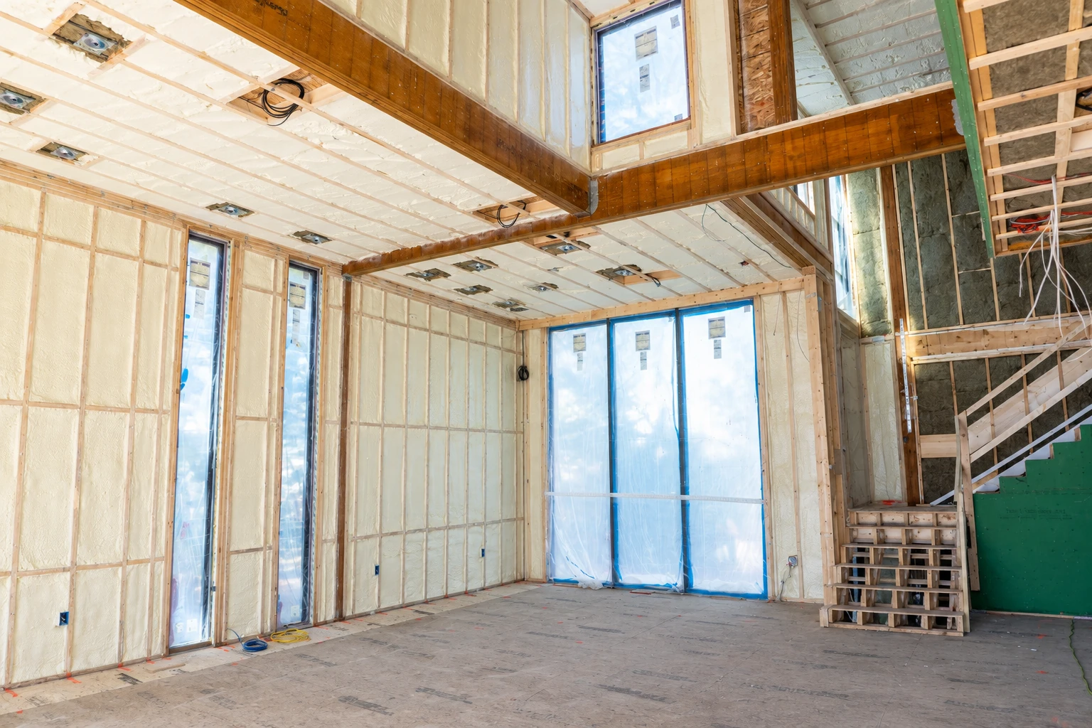 A partially finished room has pale spray foam insulation between wall studs and across the ceiling, with plastic-covered windows and exposed framing.